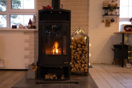 Poêle à bois noir moderne avec porte vitrée en feu et bois de chauffage rangé à côté, idéal pour un chauffage rustique et efficace.