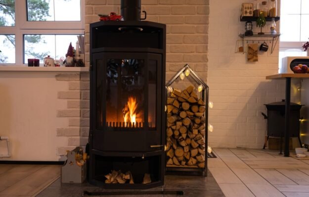 Poêle à bois noir moderne avec porte vitrée en feu et bois de chauffage rangé à côté, idéal pour un chauffage rustique et efficace.