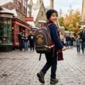 Un jeune garçon souriant porte un sac à dos Harry Potter et une écharpe de Gryffondor en marchant dans une rue pavée du village de Pré-au-Lard.